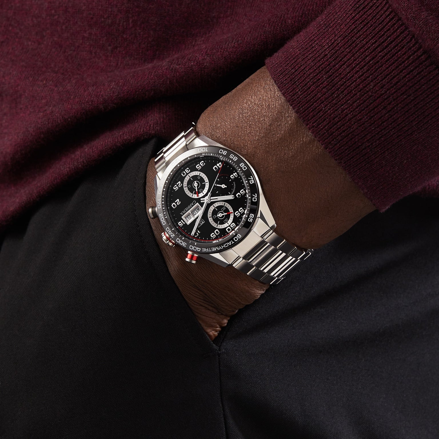 Wearing a TAG Heuer Carrera Chronograph with a silver case, black dial, and ceramic bezel, this person wears a burgundy long-sleeve shirt and black pants, with one hand in their pocket.