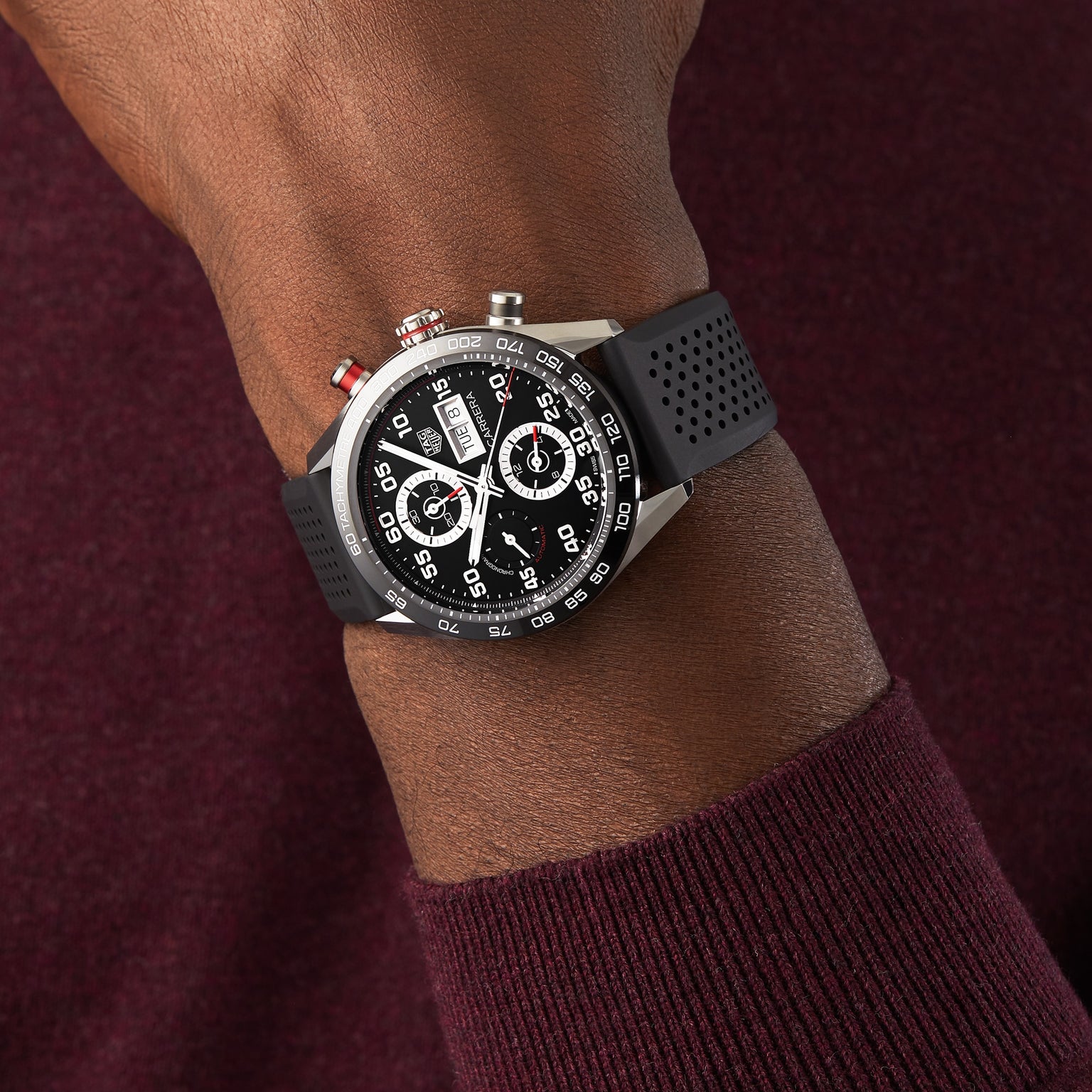 A person wearing a TAG Heuer Carrera Chronograph with a black perforated strap and calibre 16 movement, paired with a dark burgundy long-sleeve shirt.