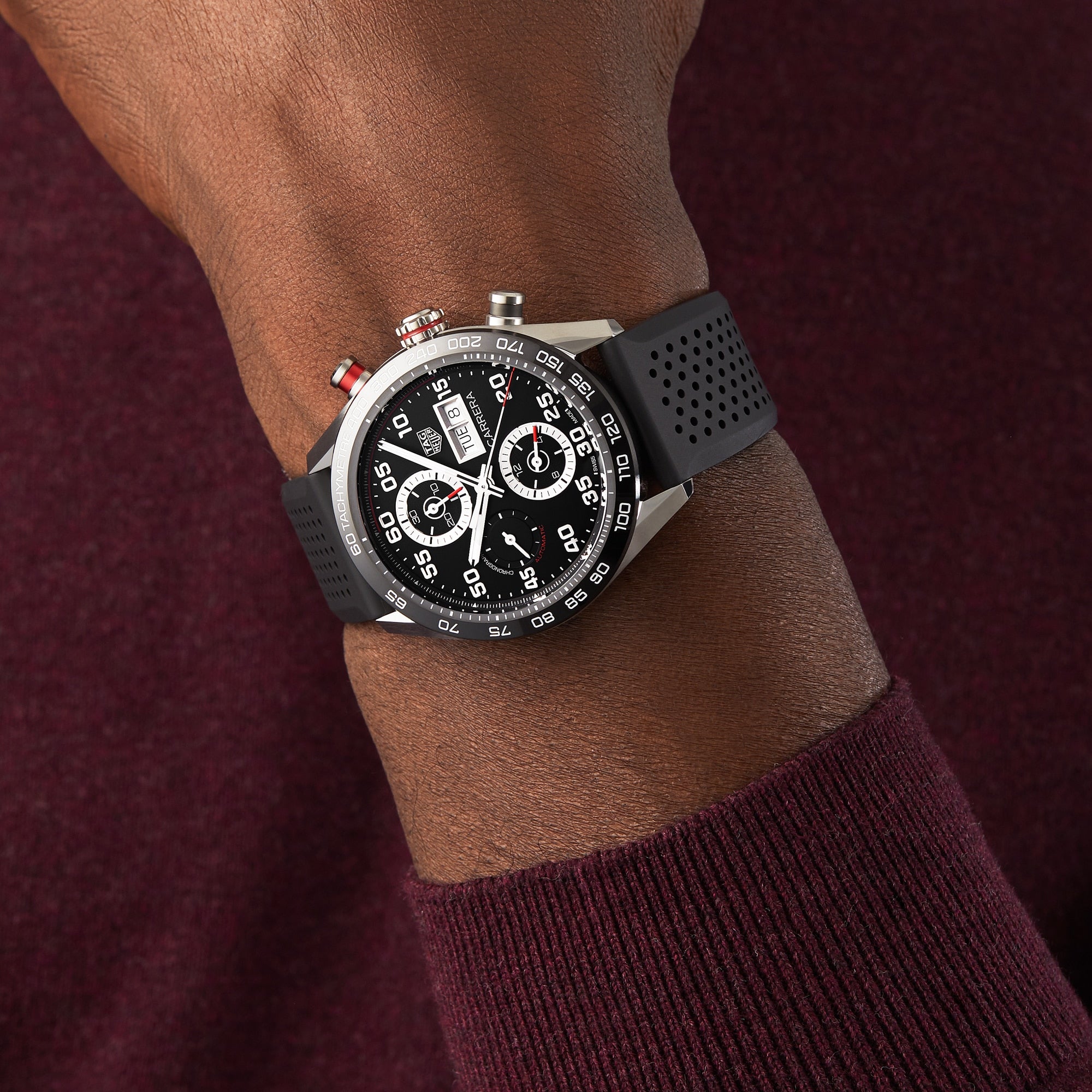A person wearing a TAG Heuer Carrera Chronograph with a black perforated strap and calibre 16 movement, paired with a dark burgundy long-sleeve shirt.