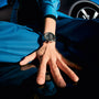A person in a blue outfit extends their hand toward the camera, showcasing a TAG Heuer Special Edition Carrera Chronograph x Porsche wristwatch; a car wheel is visible in the background.