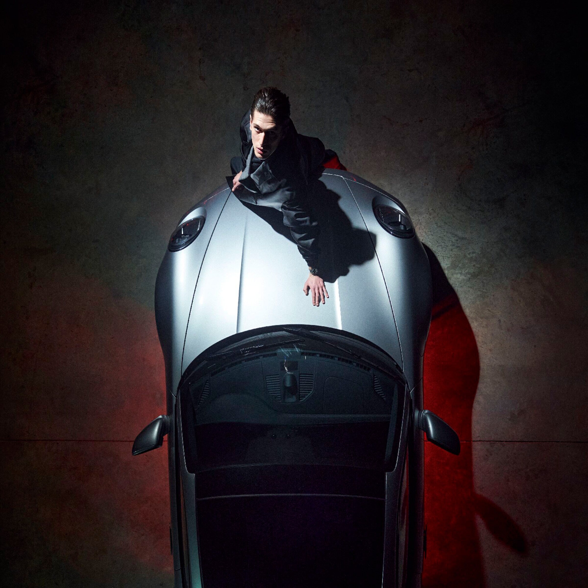 A man in dark clothing sits on the hood of a silver Porsche, viewed from above, with dramatic lighting—highlighting the TAG Heuer Special Edition Carrera Chronograph x Porsche.