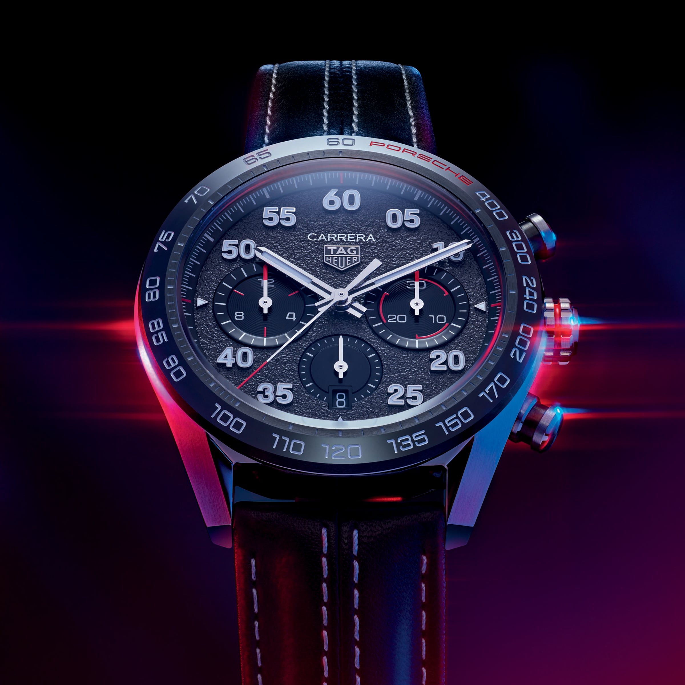 A close-up of the TAG Heuer Special Edition Carrera Chronograph x Porsche wristwatch with a black strap, dramatically lit in red and blue.
