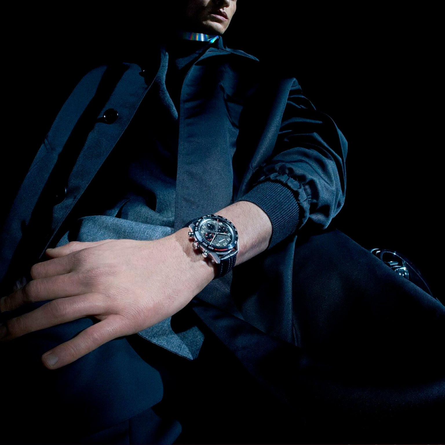 A person in dark clothing, seen from a low angle against a black background, wears the TAG Heuer Special Edition Carrera Chronograph x Porsche with a dark dial on their left wrist.