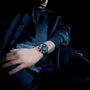 A person in dark clothing, seen from a low angle against a black background, wears the TAG Heuer Special Edition Carrera Chronograph x Porsche with a dark dial on their left wrist.