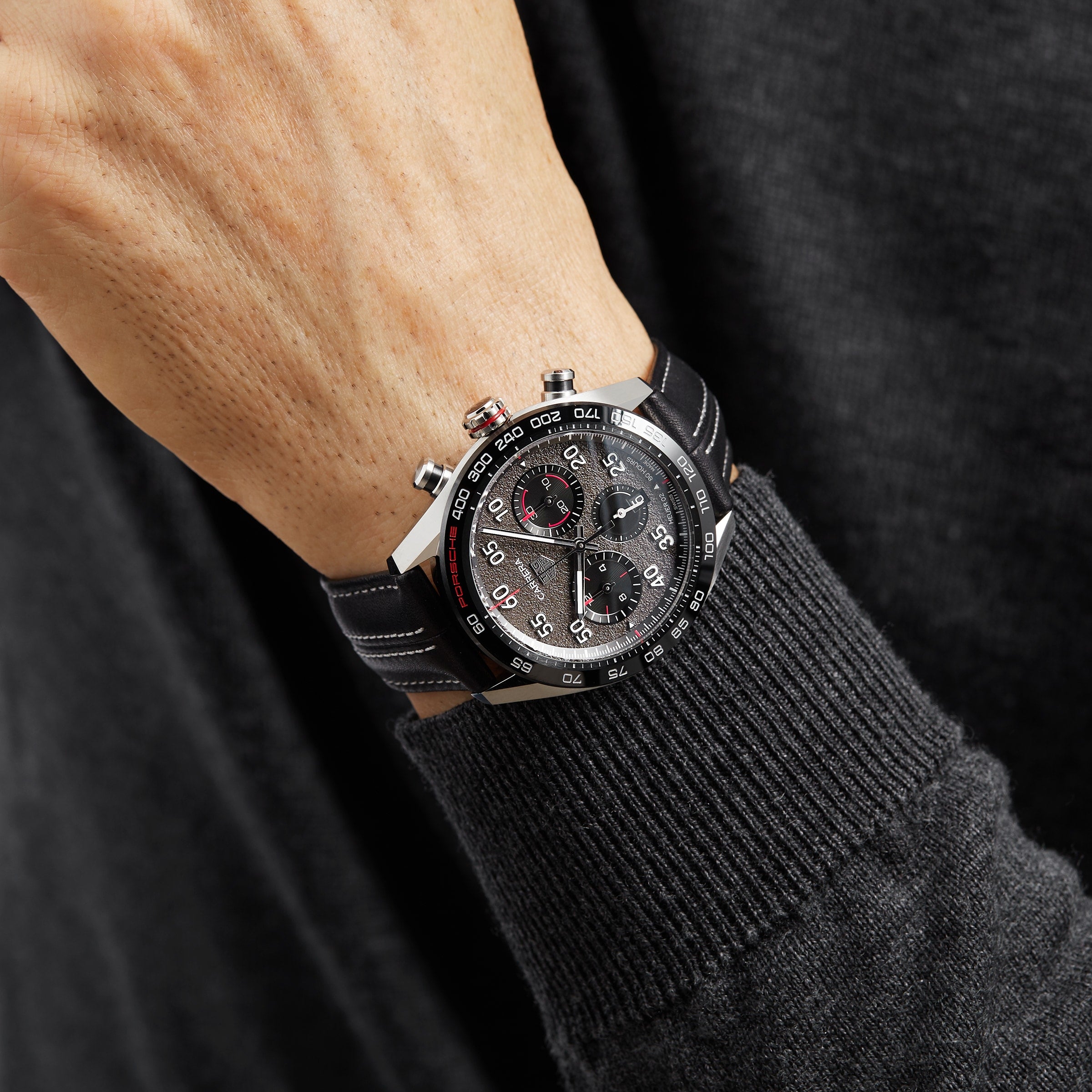 A person wearing a TAG Heuer Special Edition Carrera Chronograph x Porsche with a black leather strap and racing-style dial, dressed in a dark sweater.