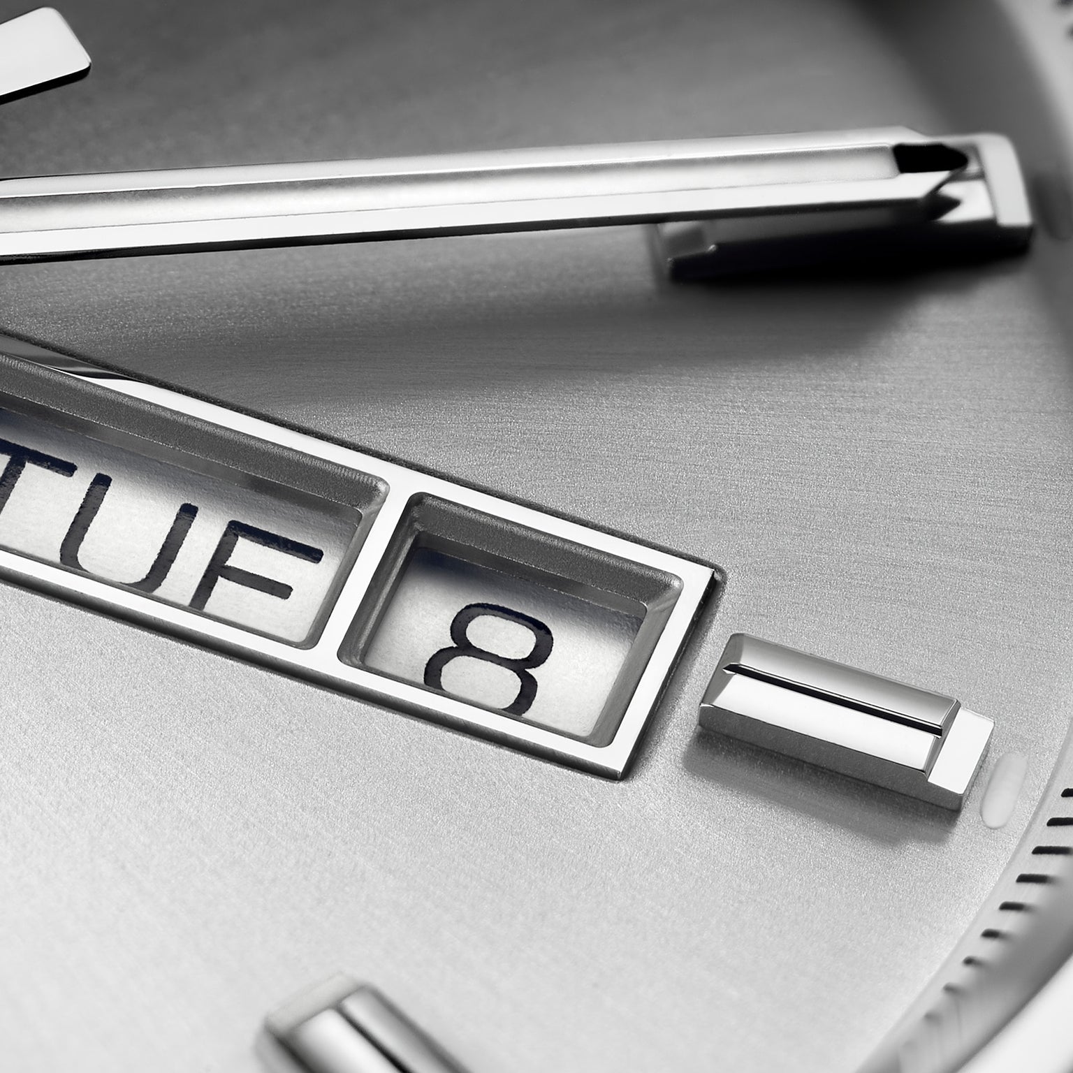 Close-up of the TAG Heuer Carrera Day-Date watch face, showing metallic hour markers, calibre 5 movement, and a partial day-date window with TUE 8 on a brushed silver background.