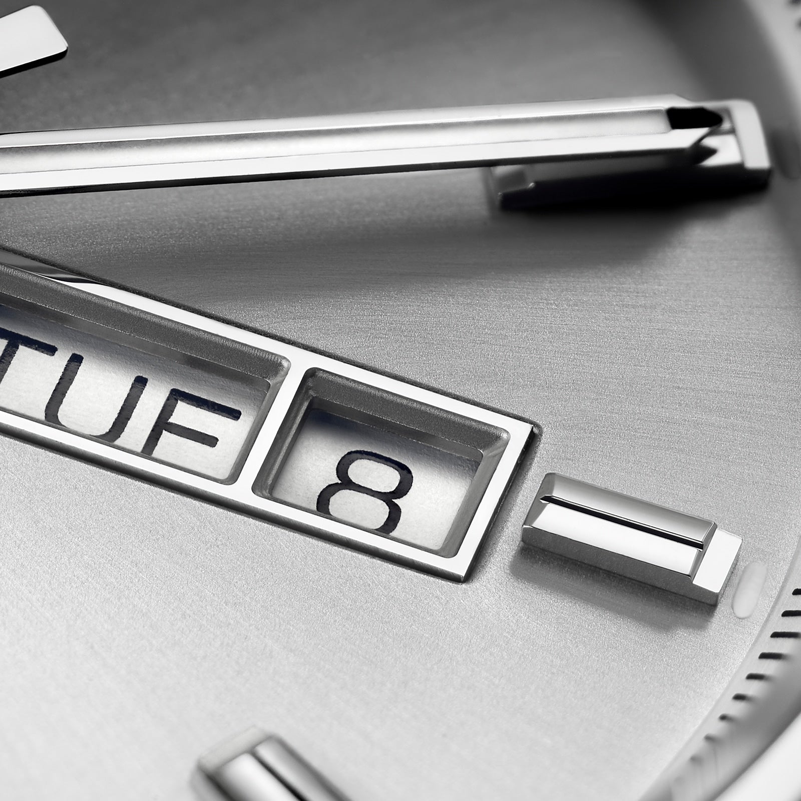 Close-up of the TAG Heuer Carrera Day-Date watch face, showing metallic hour markers, calibre 5 movement, and a partial day-date window with TUE 8 on a brushed silver background.