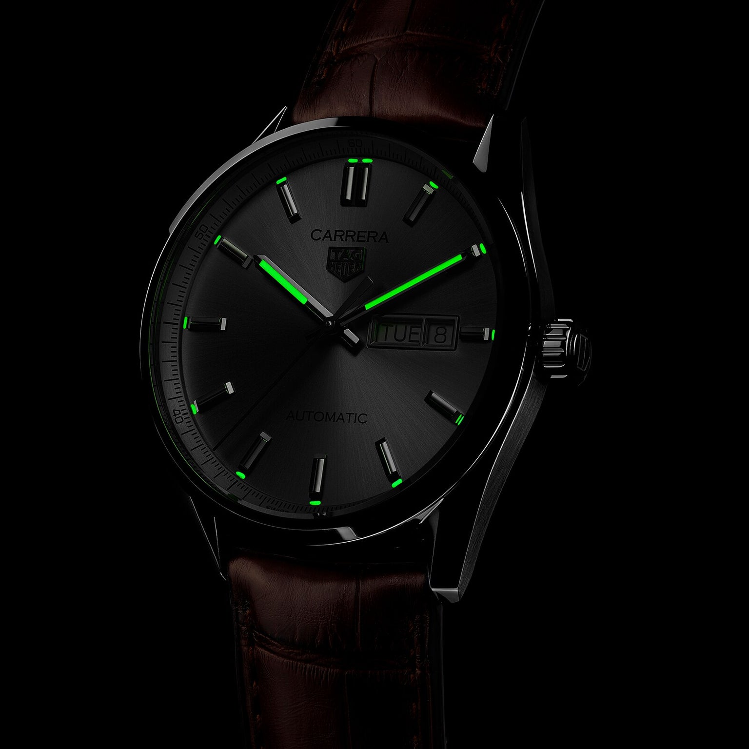 The TAG Heuer Carrera Day-Date features a brown alligator leather strap, black dial with luminous green markers and hands, Calibre 5 movement, and displays both day and date.