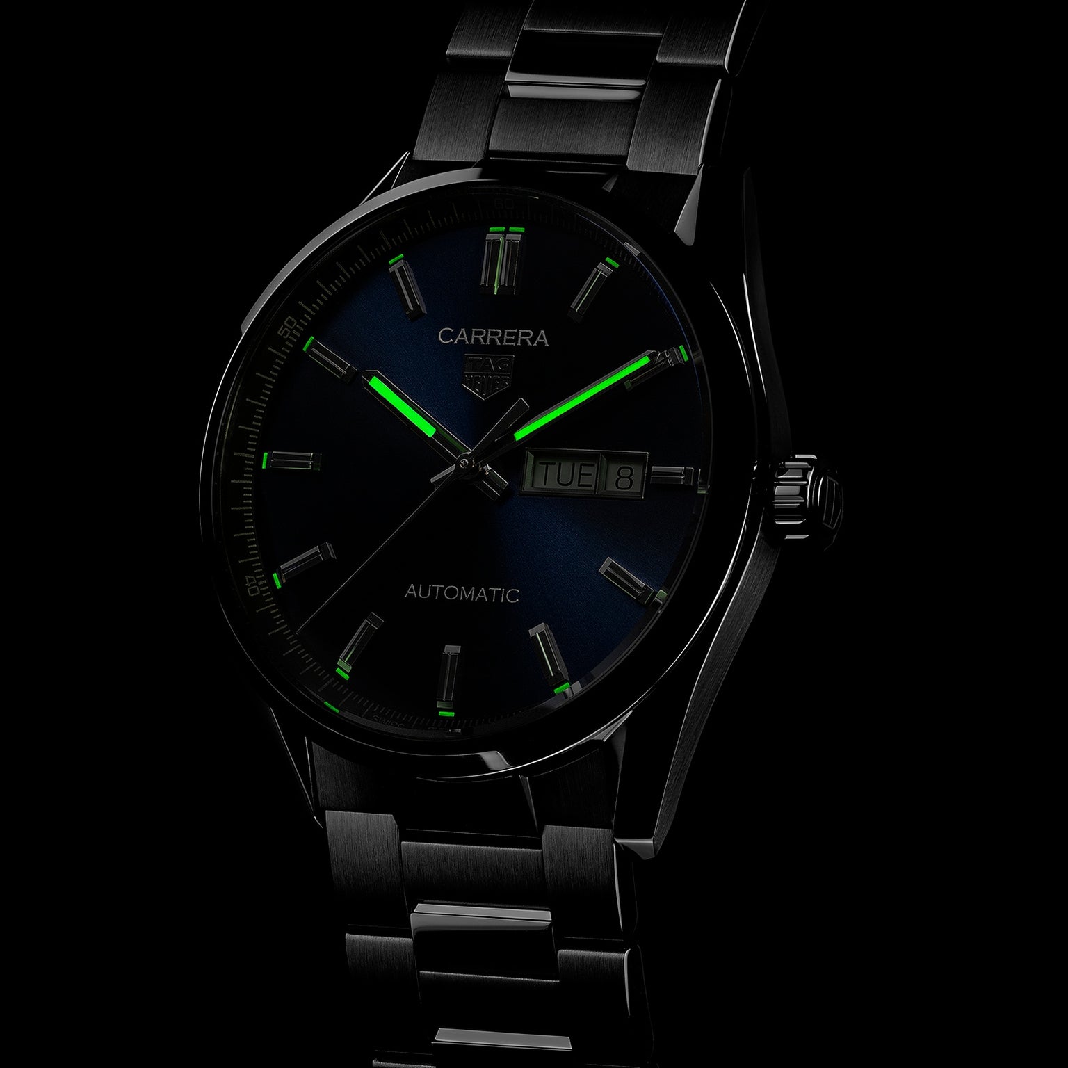 A black TAG Heuer Carrera Day-Date watch with luminescent green markers, calibre 5 automatic movement, and a day-date display reading Tuesday 8 is shown against a dark background.