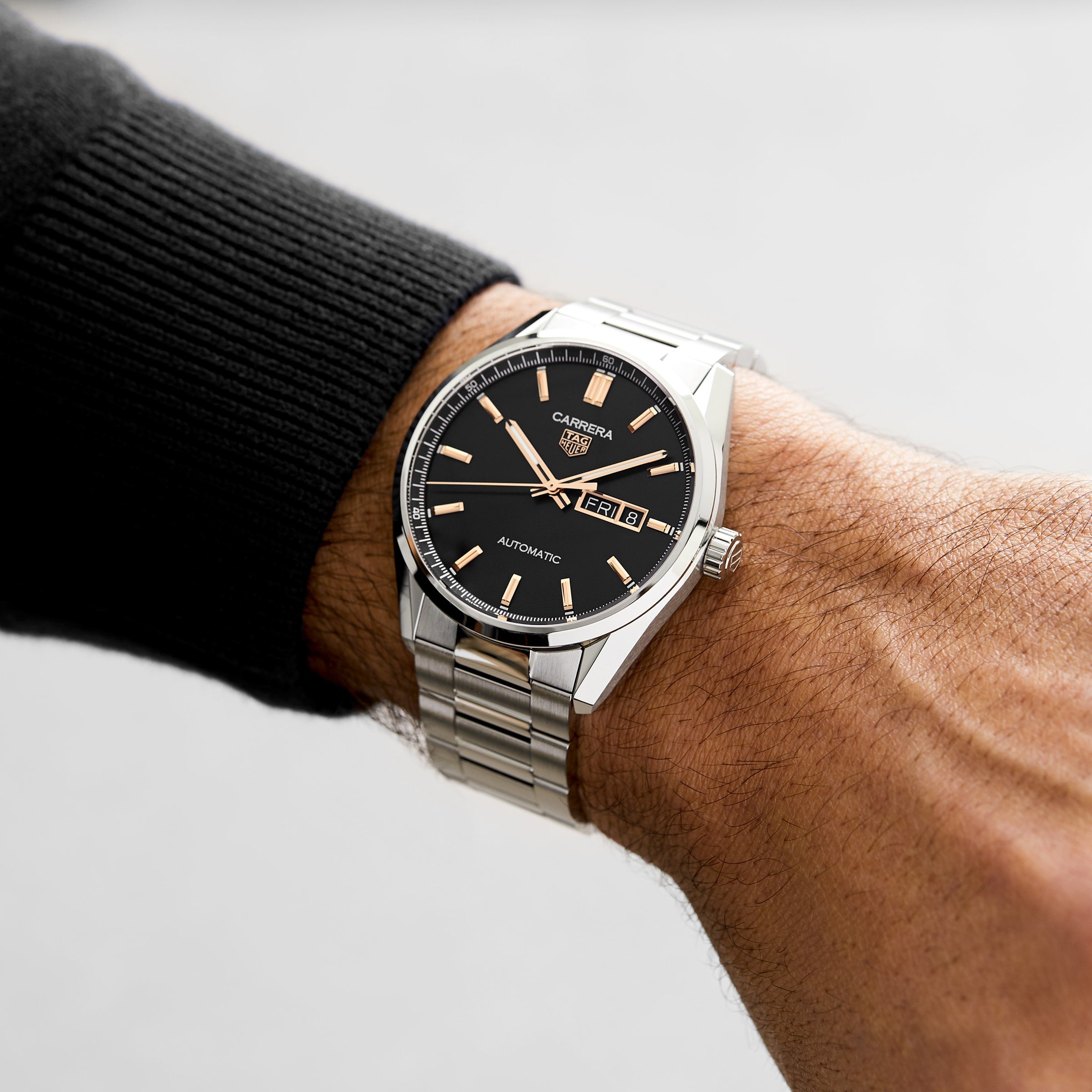 A close-up of a person wearing a silver TAG Heuer Carrera Day-Date on their left wrist, showcasing a black dial and metal bracelet—an enduring symbol of precision.
