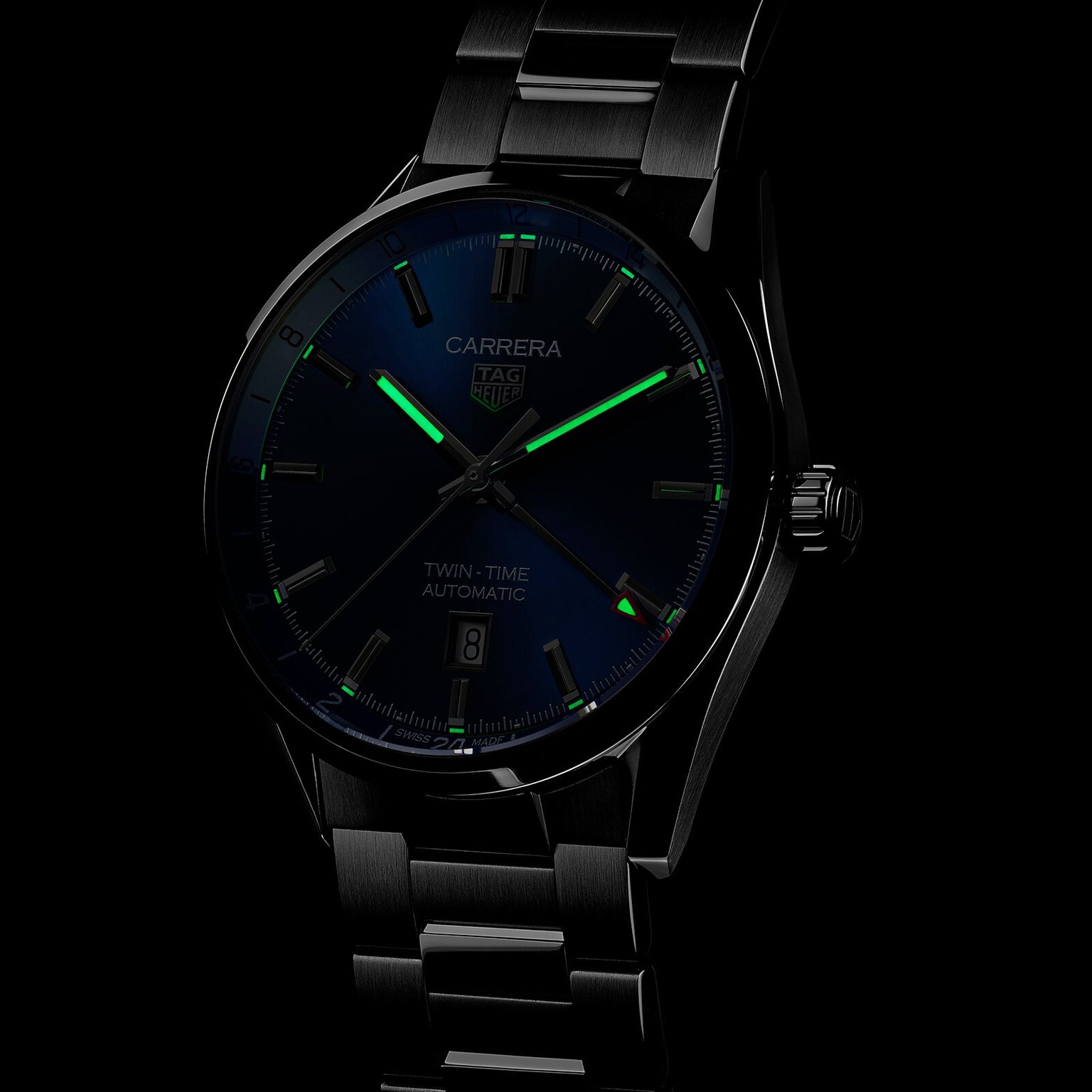 The TAG Heuer Carrera Twin-Time watch features GMT functionality, a dark dial with luminous green hands, and a stainless steel bracelet, shown against a black background.