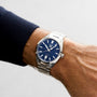 A person wearing a TAG Heuer Carrera Date watch with a stainless steel case, blue dial, date display, and automatic movement on their left wrist.