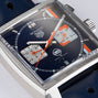Close-up of the TAG Heuer Special Edition Monaco Gulf wristwatch with a square blue dial, orange Gulf stripe and logo, two subdials, blue leather strap, and stainless steel case.