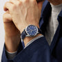 Wearing a TAG Heuer Carrera Date with a blue face and blue leather strap, powered by the calibre 5 automatic movement, a person in a blue jacket and grey shirt rests their chin on their hands—embodying endurance and performance.