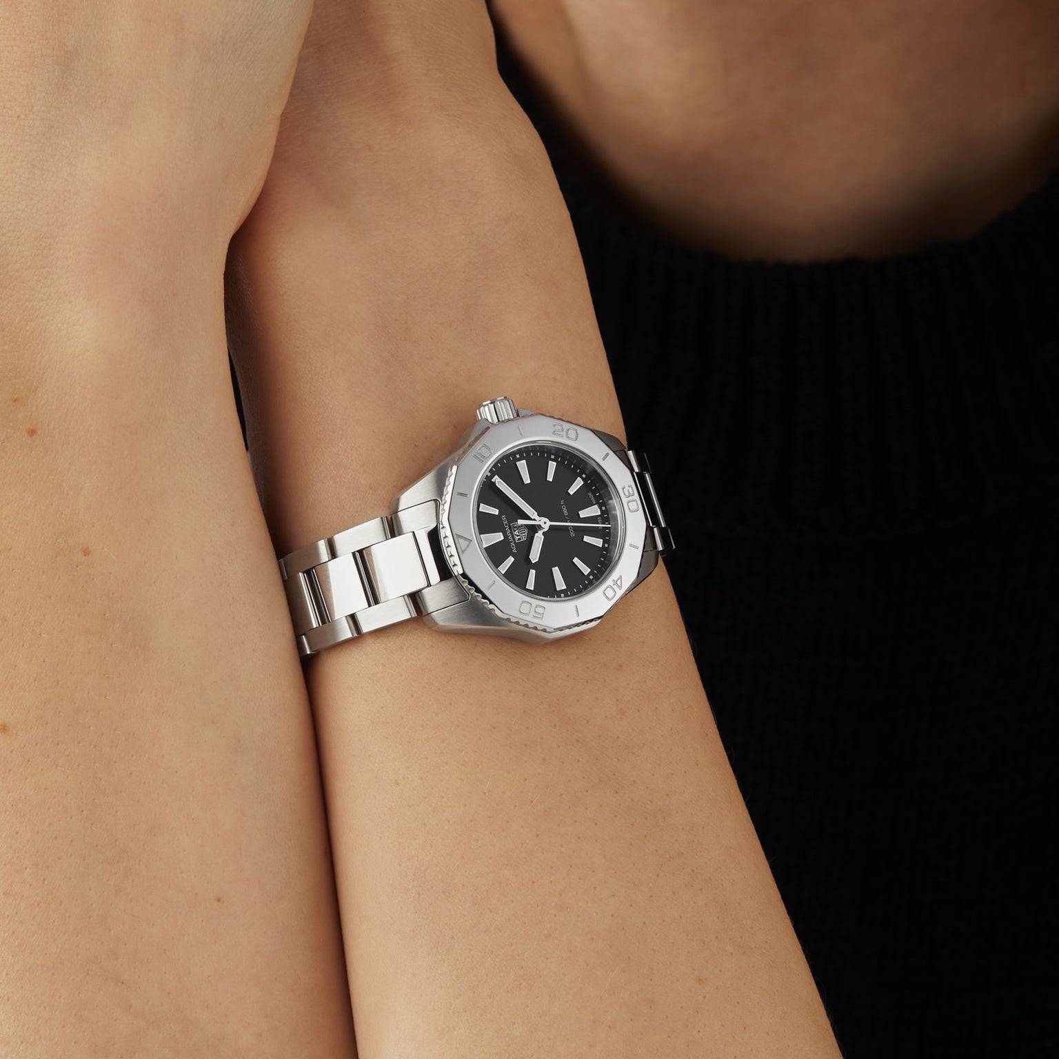 A person with arms crossed against a black top wears a TAG Heuer Aquaracer Professional 200 sports watch, featuring a stainless steel case, black dial, and silver indices, on their left wrist.