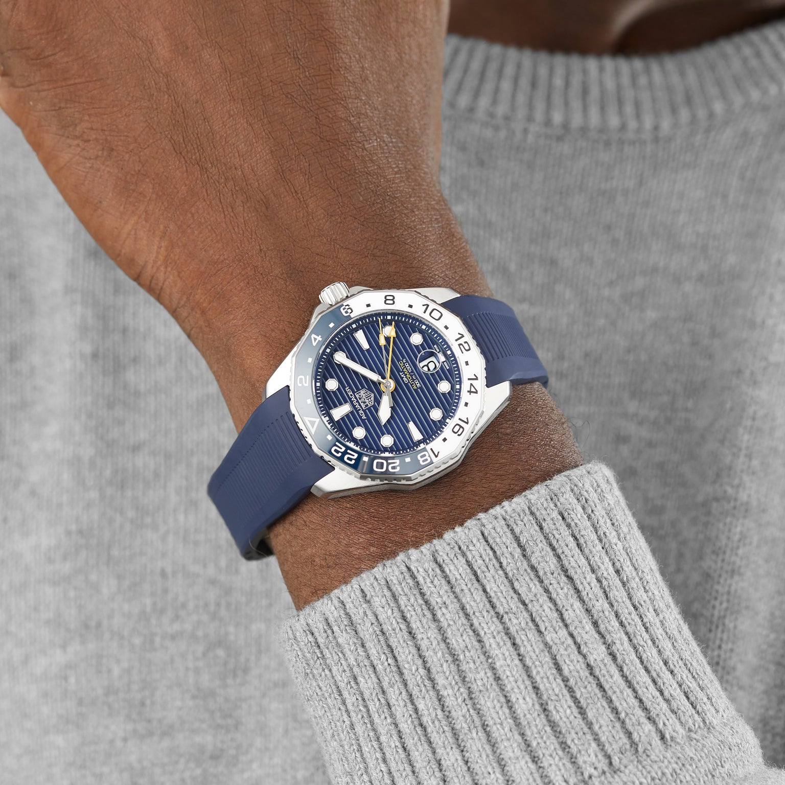 A close-up of a person wearing the TAG Heuer Aquaracer Professional 300 GMT with a blue strap and patterned blue dial, paired with a light grey knit sweater.