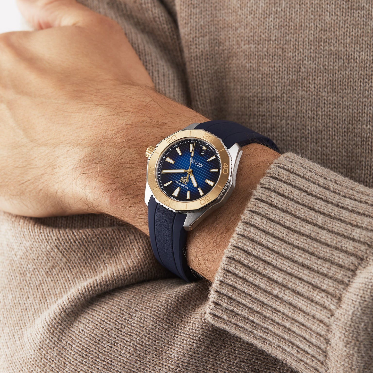 A person wears a TAG Heuer Aquaracer Professional 200 Date, a striking gold and blue sports watch, over a beige knit sweater.