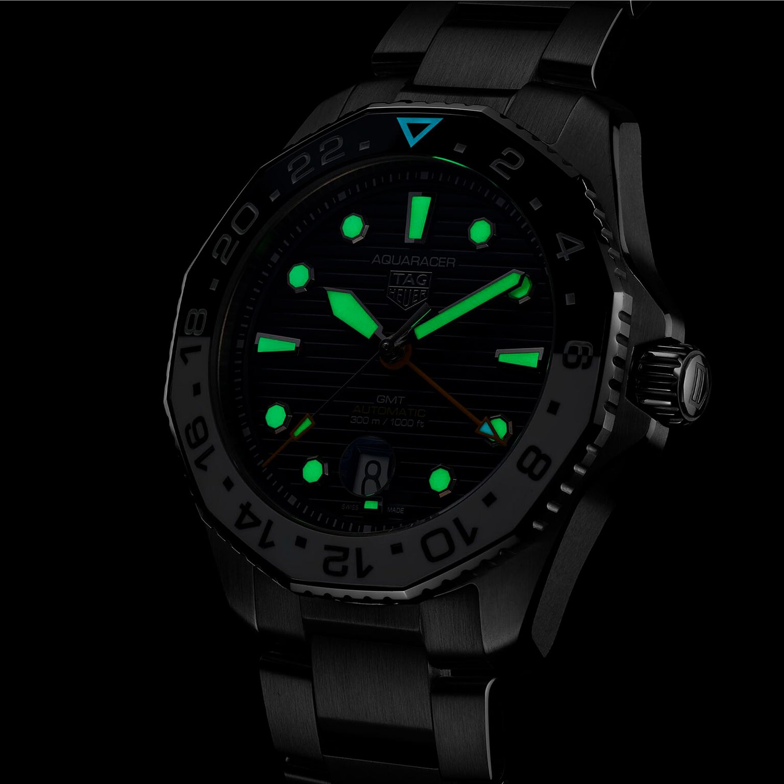 The TAG Heuer Aquaracer Professional 300 GMT sports watch features a black metal band and case, GMT movement, and luminous green hour markers, hands, and indices that glow in the dark.