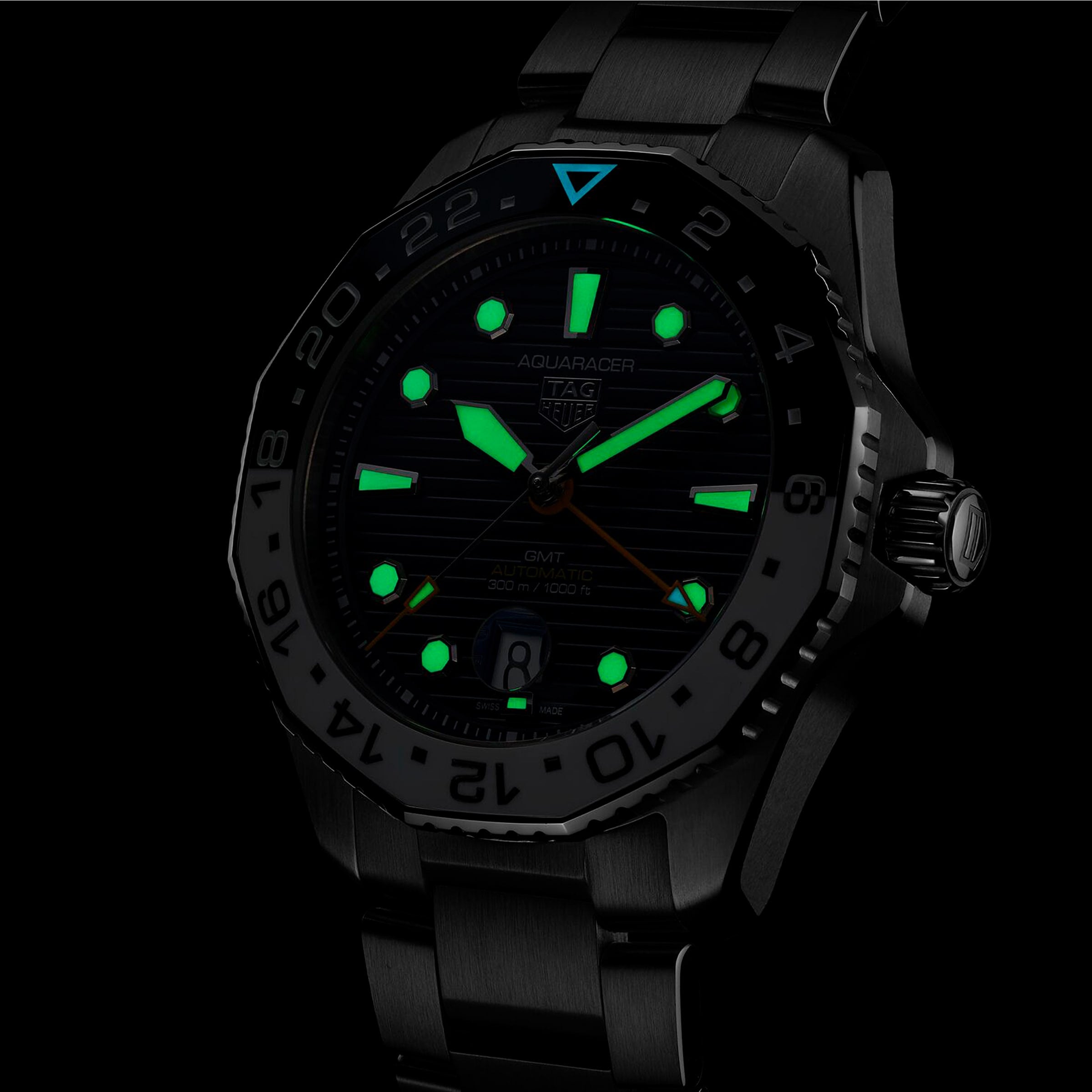 The TAG Heuer Aquaracer Professional 300 GMT sports watch features a black metal band and case, GMT movement, and luminous green hour markers, hands, and indices that glow in the dark.