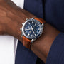 A TAG Heuer Carrera Chronograph with a brown leather strap and black dial is worn on a wrist beside a navy blue sleeve.