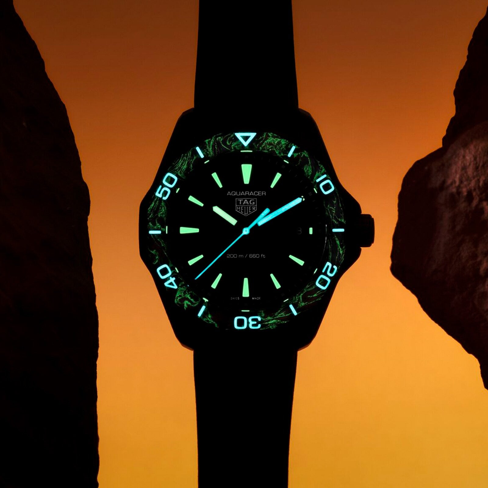A TAG Heuer Aquaracer Professional 200 Solargraph watch with luminous markers and hands is showcased against an orange backdrop, set between two dark rocks.