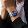 A person in a white shirt and black jacket wears a TAG Heuer Special Edition Formula 1 Chronograph X Gulf wristwatch, featuring a blue face and multiple dials.
