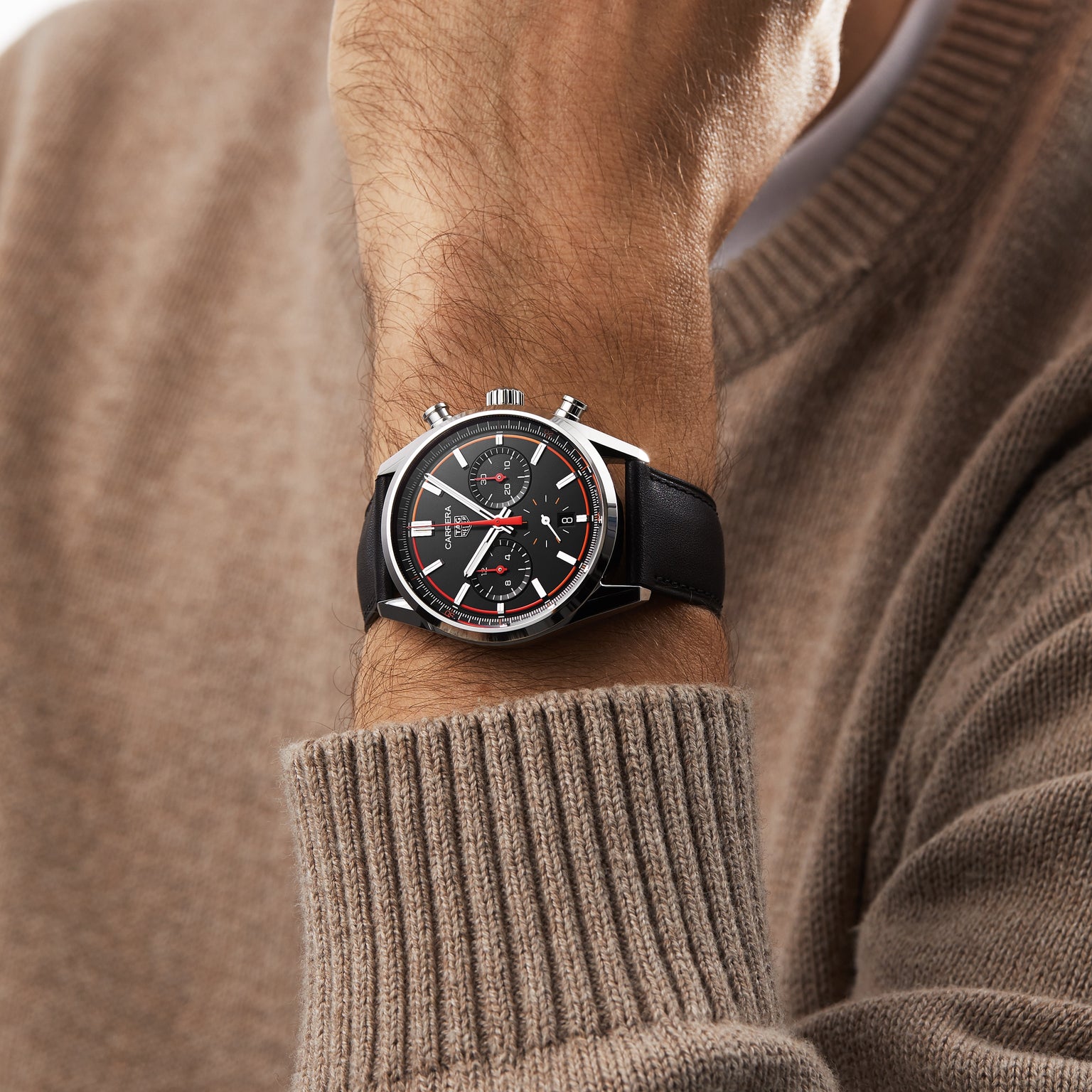 A person in a beige sweater showcases the TAG Heuer Carrera Chronograph, which features a black dial, chronograph function, and is powered by the Calibre HEUER 02 movement.