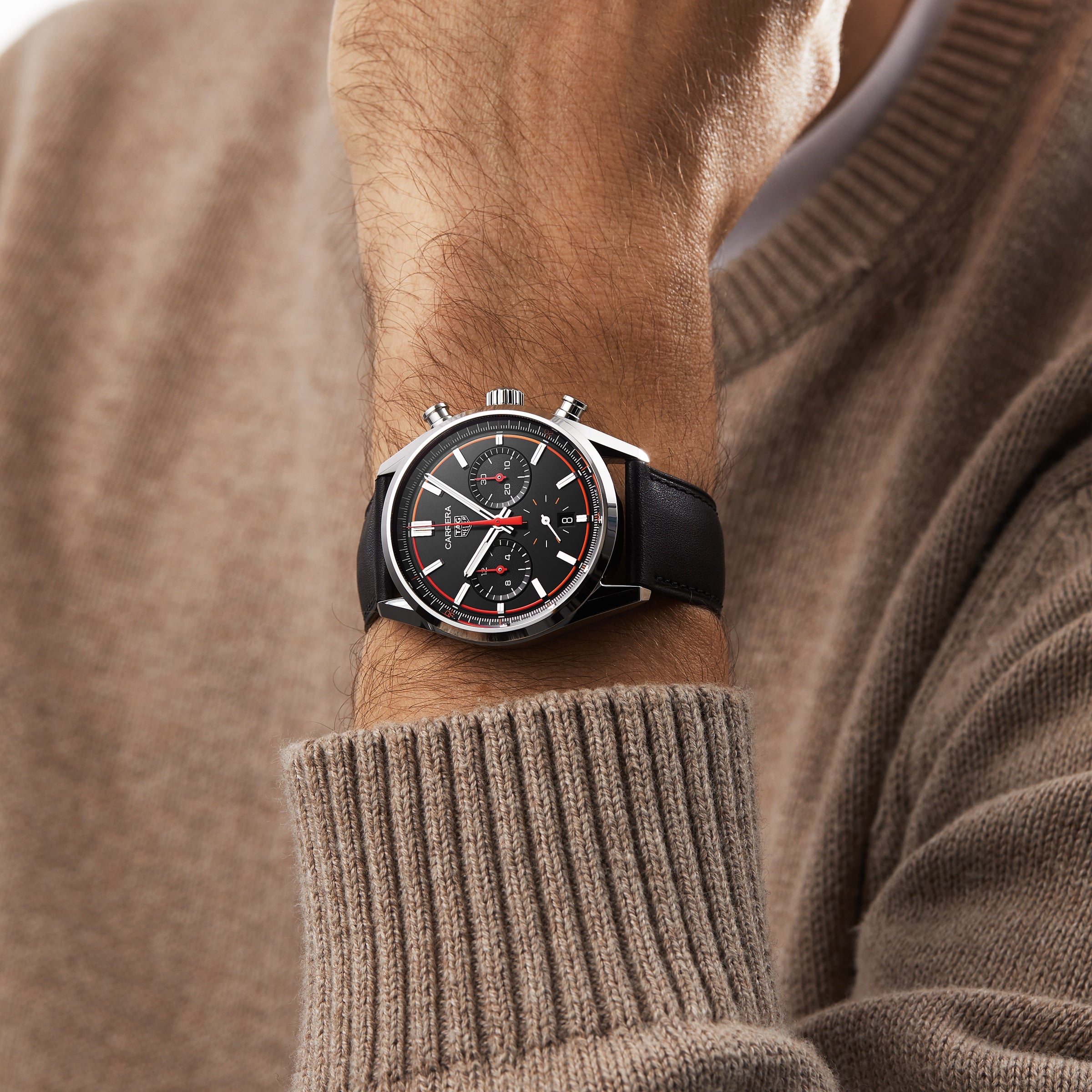 A person in a beige sweater showcases the TAG Heuer Carrera Chronograph, which features a black dial, chronograph function, and is powered by the Calibre HEUER 02 movement.