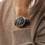 A person in a beige sweater showcases the TAG Heuer Carrera Chronograph, which features a black dial, chronograph function, and is powered by the Calibre HEUER 02 movement.