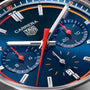 Close-up of a blue TAG Heuer Carrera Chronograph watch face, featuring red and white hands, two subdials, and a date window showing the number 8.