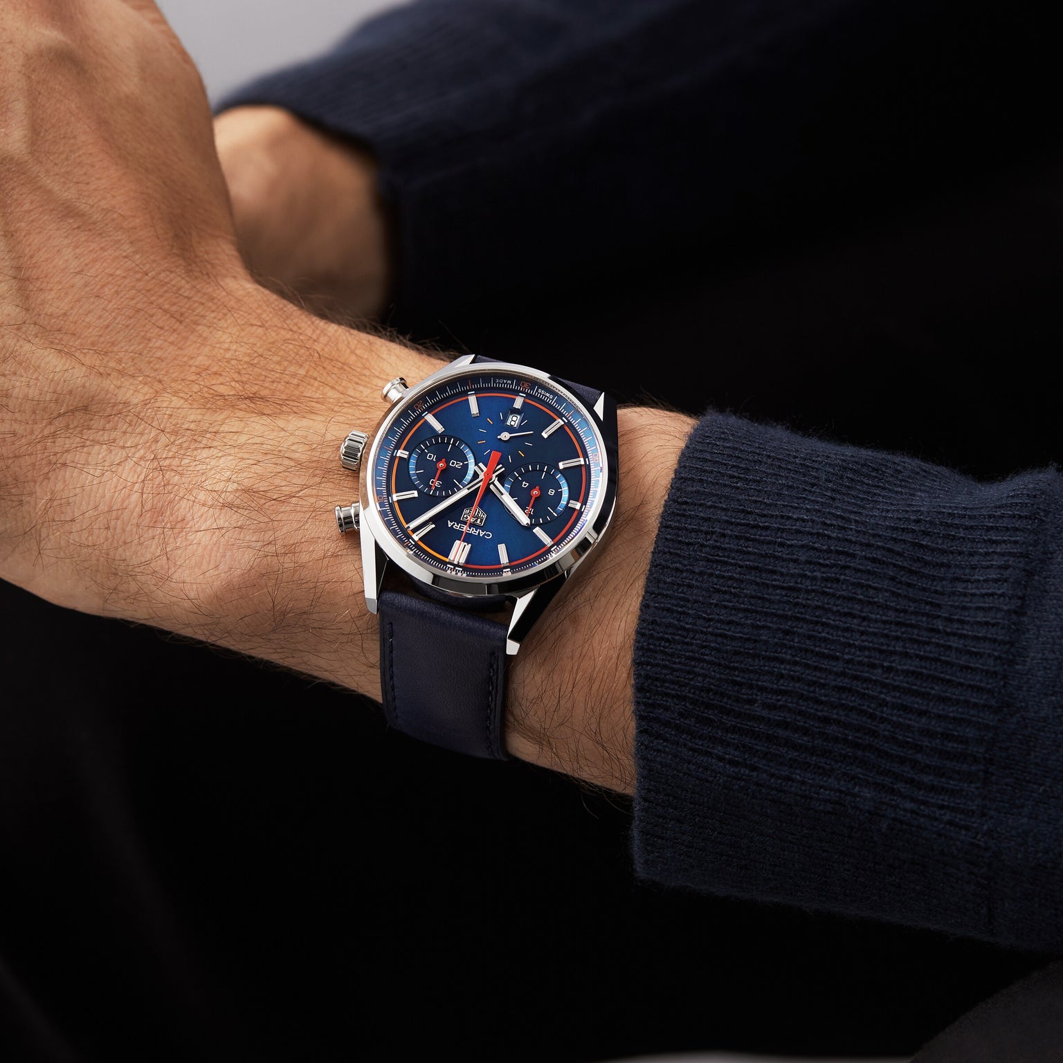 A person wearing a TAG Heuer Carrera Chronograph with a navy dial and silver case, paired with a navy long sleeve sweater.