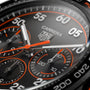 Close-up of the TAG Heuer Special Edition Carrera Chronograph x Porsche Orange Racing watch face, showcasing chronograph dials, orange accents, and bold white numerals on a brushed metal background.