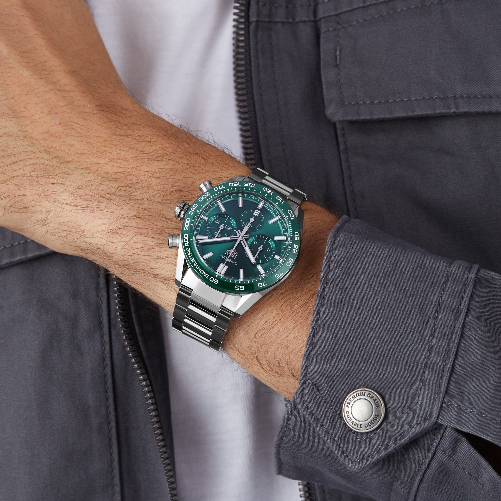 A person wearing a dark gray jacket and a TAG Heuer Carrera Chronograph wristwatch with a green dial and three subdials, featuring the Heuer 02 calibre inspired by motor-racing heritage, on their left wrist.