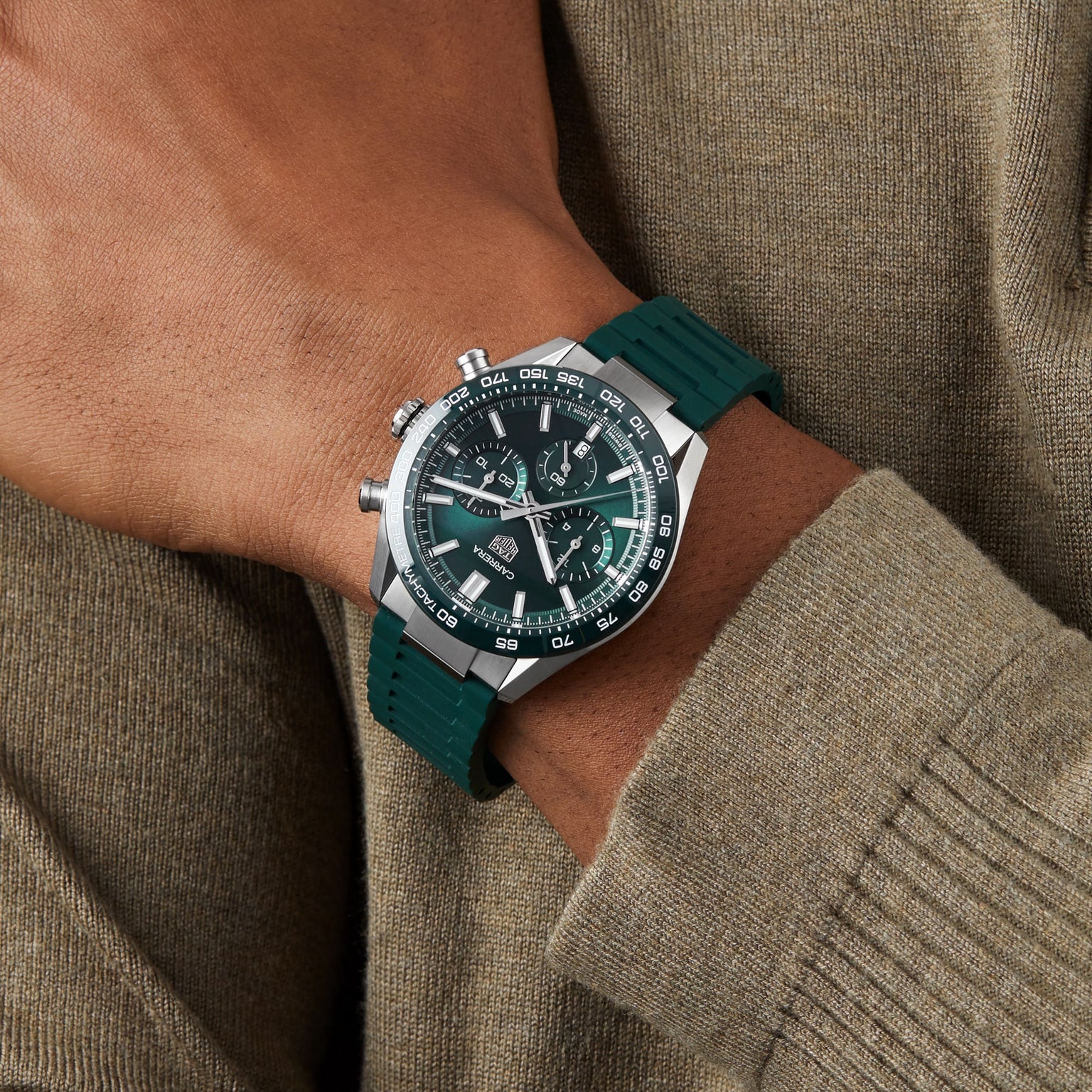 Someone sports a TAG Heuer Carrera Chronograph with a green rubber strap, green dial, and silver case, styled with a khaki long-sleeve shirt—a classic tribute to TAG Heuers motor-racing legacy.