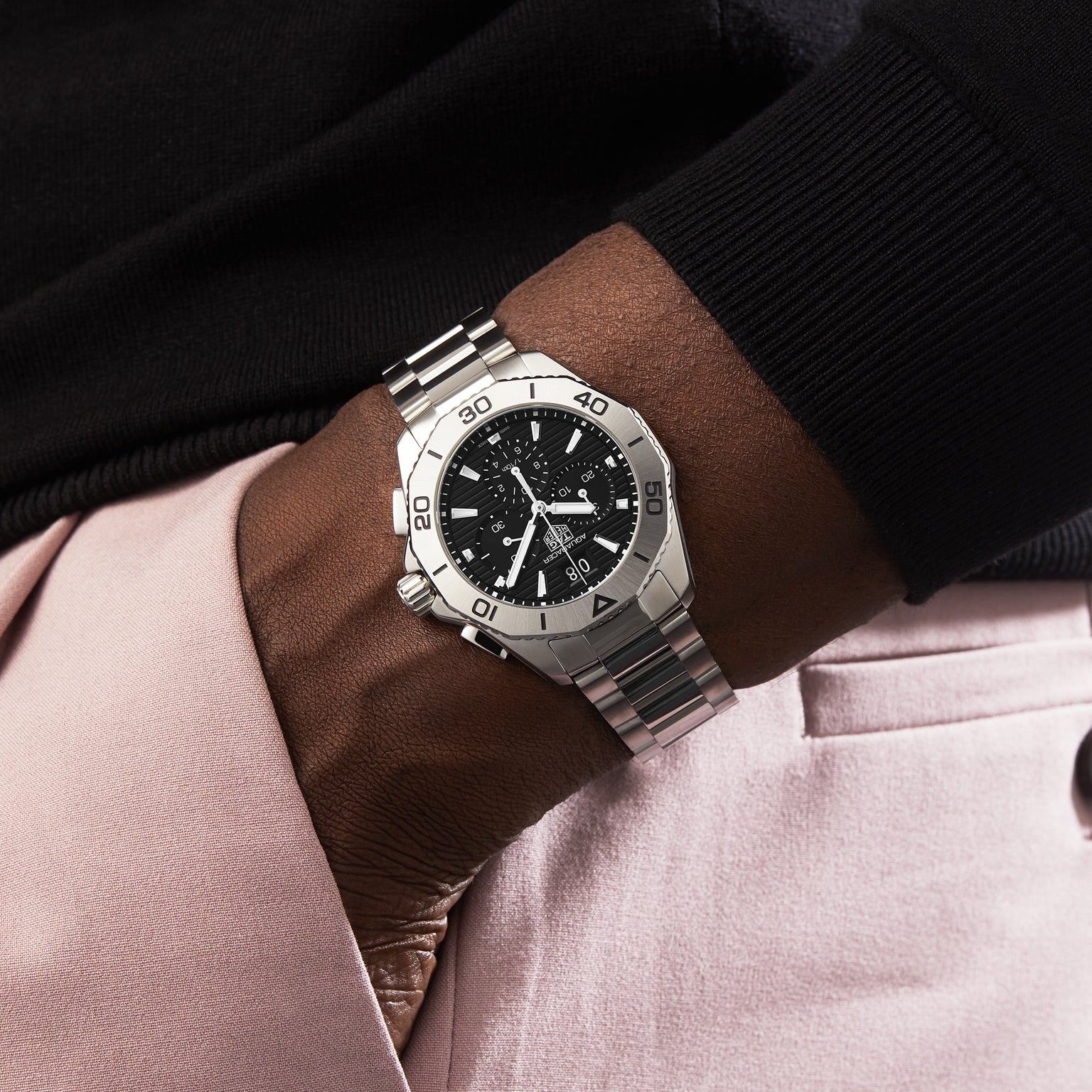 A person wearing a TAG Heuer Aquaracer Professional 200 Chronograph with a black dial and metal band rests their hand in the pocket of light pink pants.