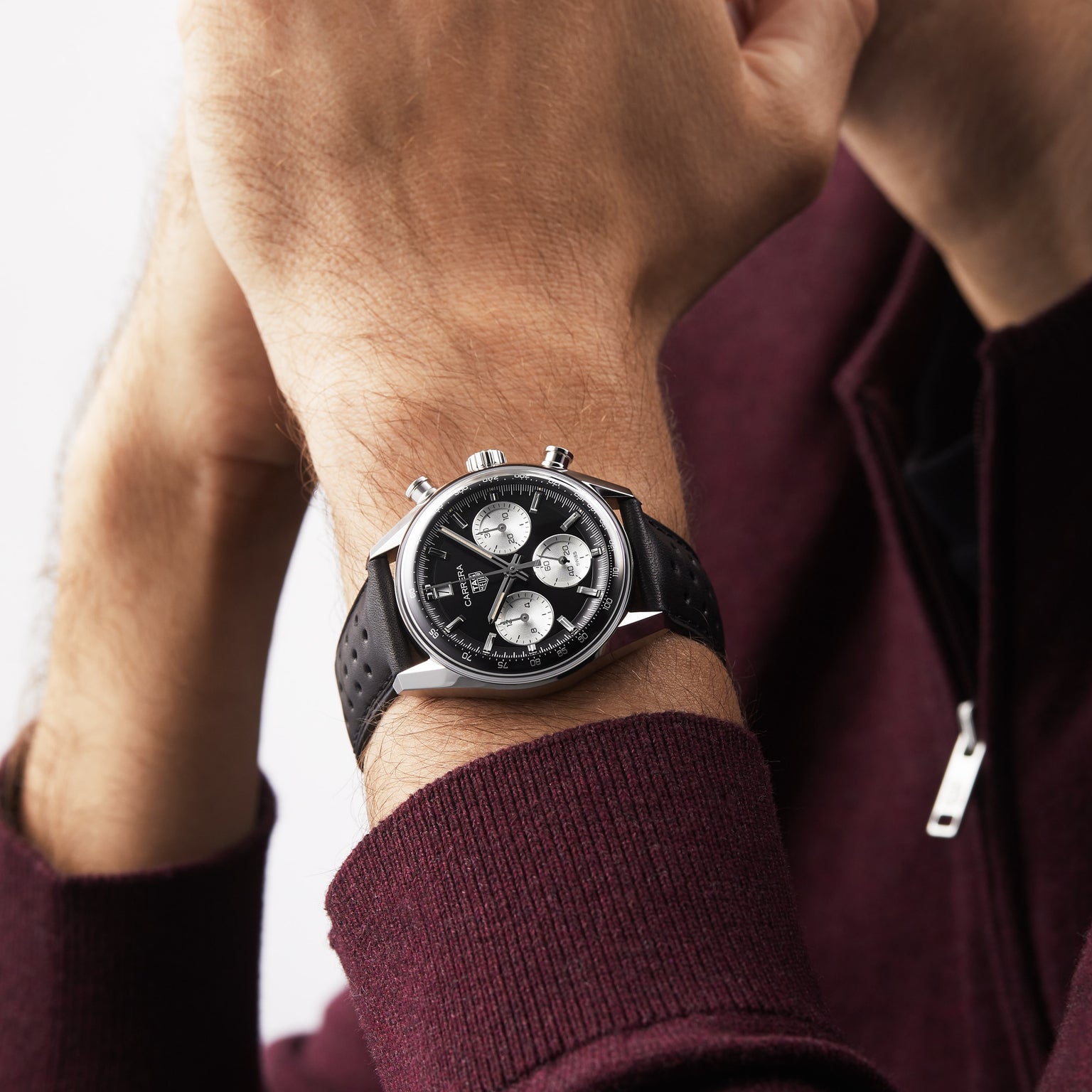 A person in a maroon zip-up sweater wears a TAG Heuer Carrera Chronograph with a black leather strap, silver case, and three subdials on their left wrist.