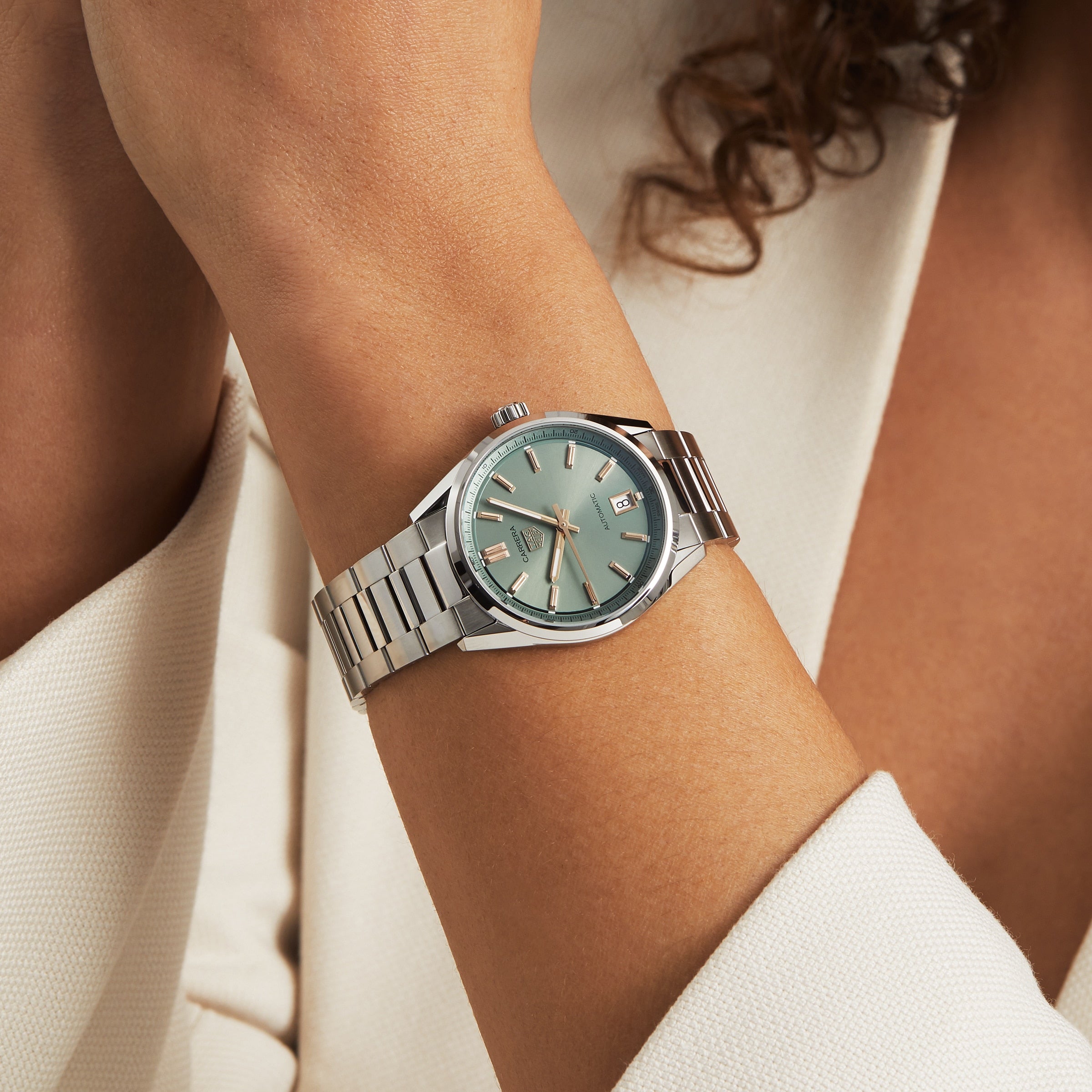 A person dressed in a cream blazer showcases the elegance of the TAG Heuer Carrera Date, a silver wristwatch featuring a light green face and date display.