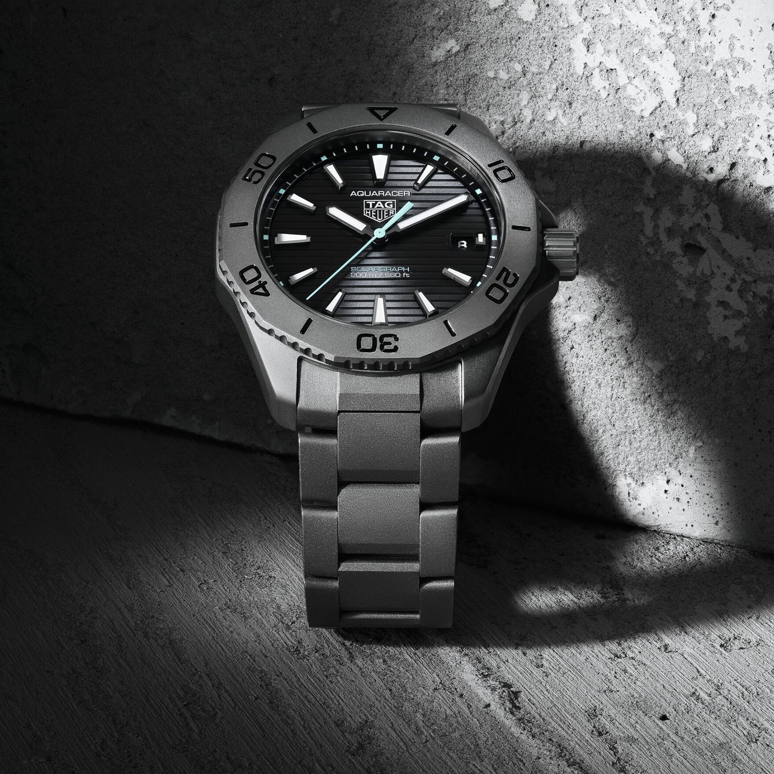 A TAG Heuer Aquaracer Professional 200 Solargraph wristwatch with a metal band sits on a textured concrete surface, partially lit to highlight the elegance of this solar-powered timepiece.