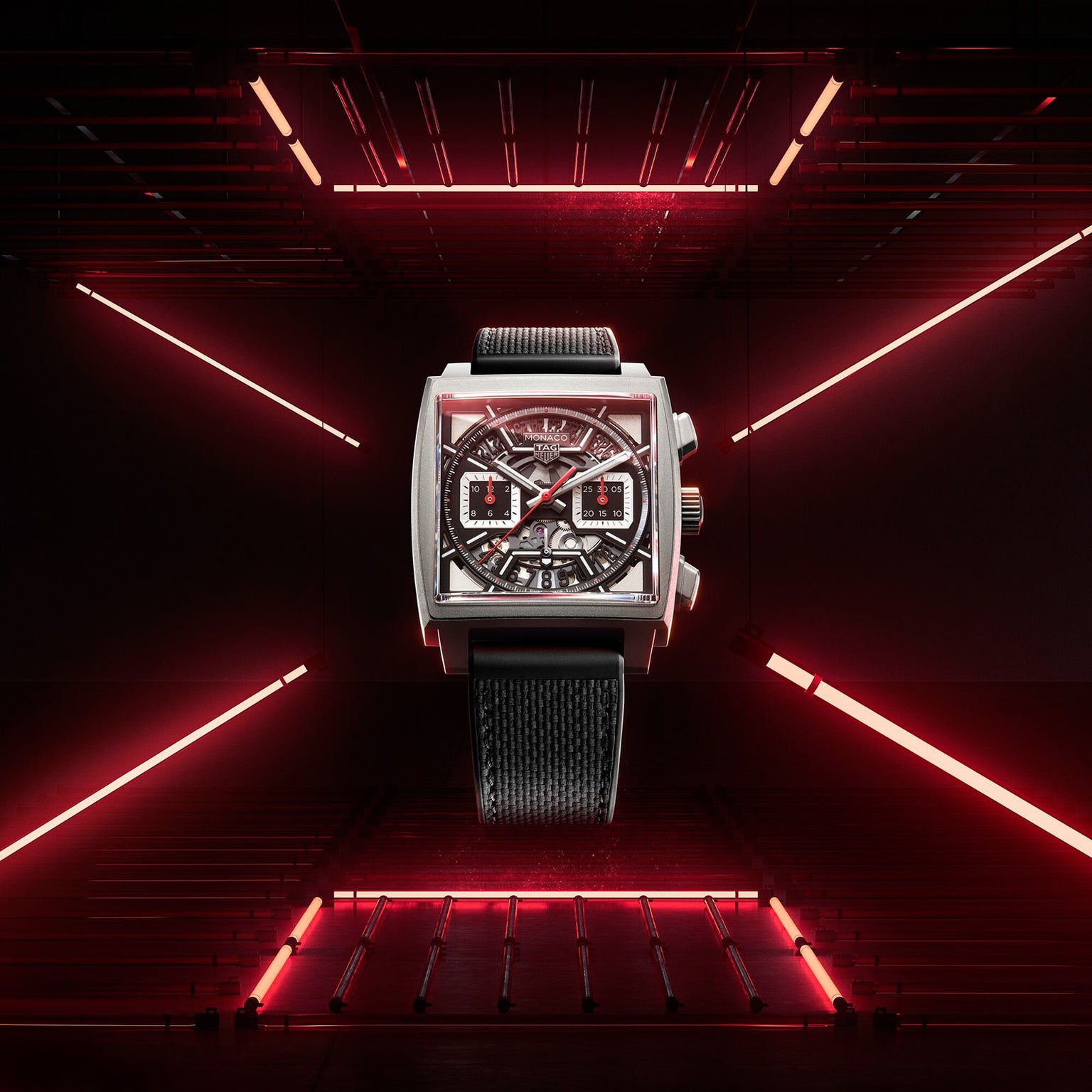The TAG Heuer Monaco Chronograph wristwatch, featuring a square face, black strap, and skeletonised dial with calibre Heuer 02 movement, is displayed against a dark background illuminated by red neon lights.