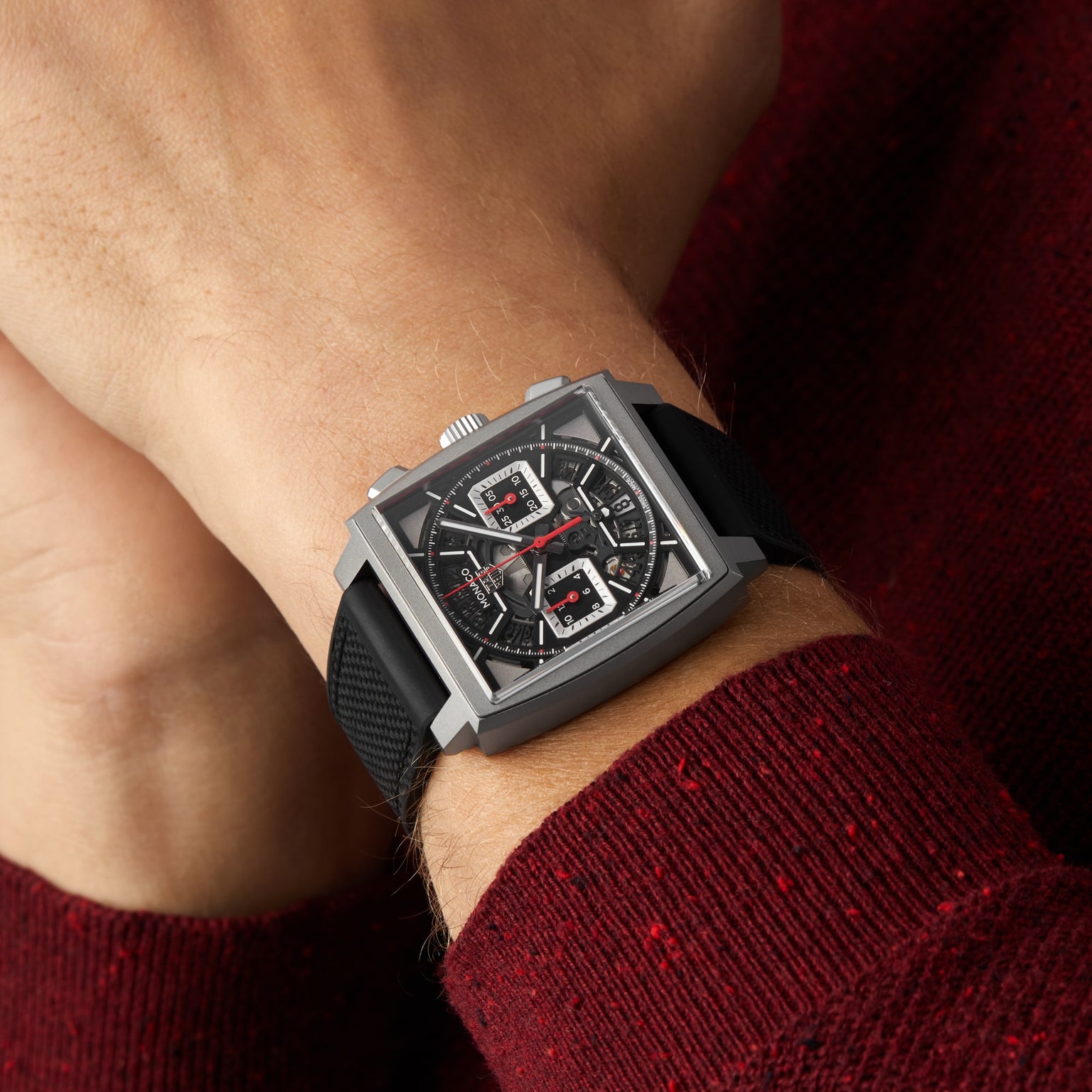 A person wearing a TAG Heuer Monaco Chronograph with a black strap and skeletonised dial rests their hands together on a maroon sweater.