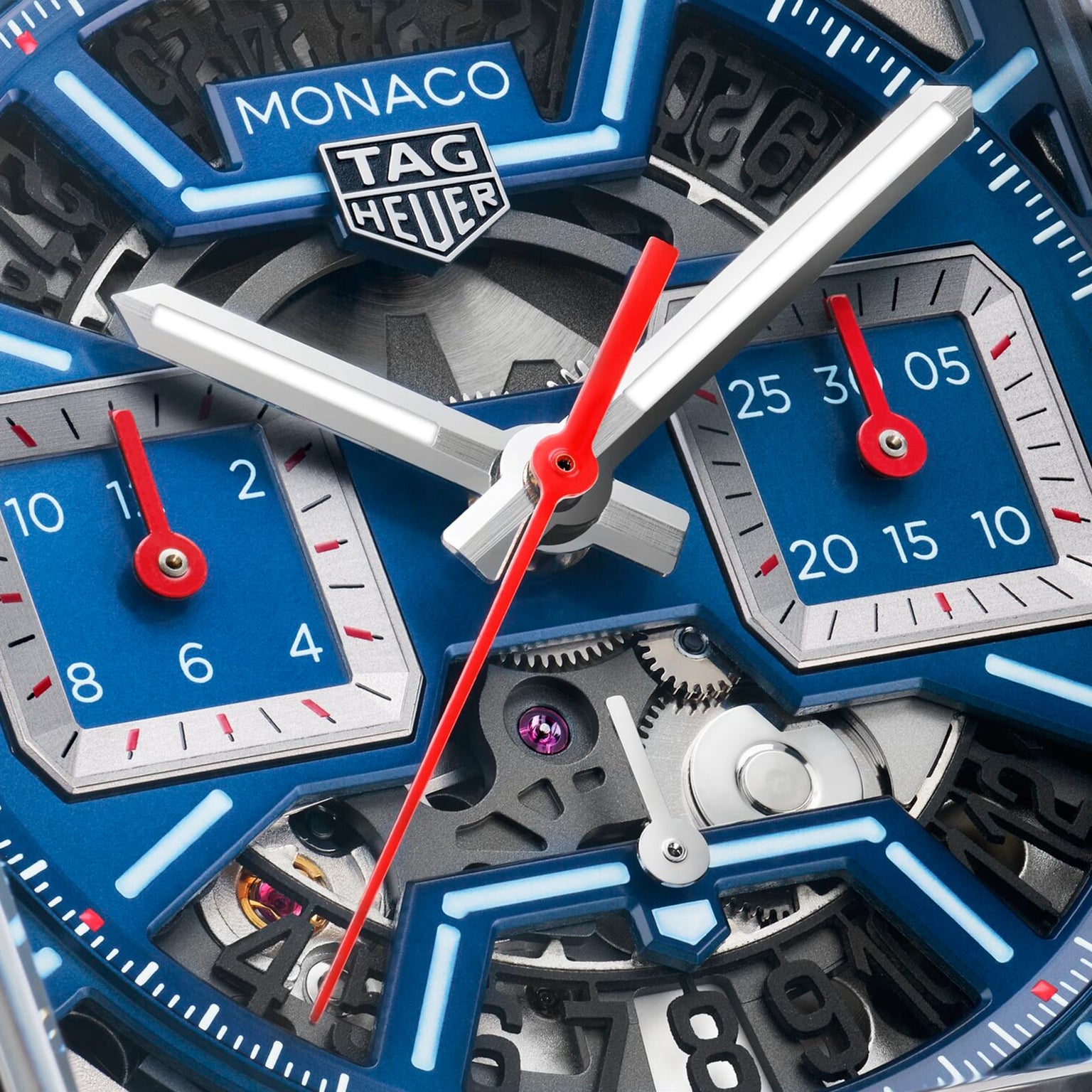Close-up of a TAG Heuer Monaco Chronograph watch face with a skeletonized dial, blue subdials, white hour and minute hands, red second hand, and visible Heuer 02 mechanical movement.