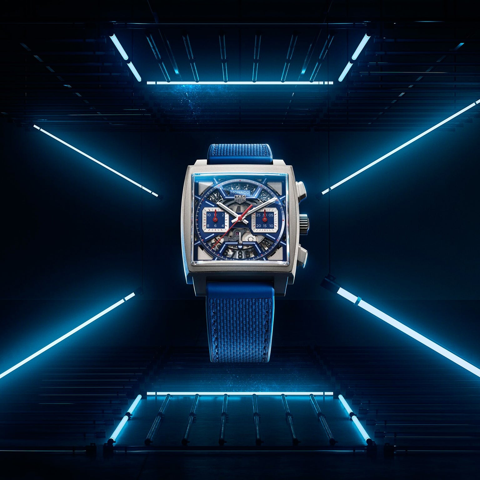 A TAG Heuer Monaco Chronograph wristwatch with a blue strap and silver square case, featuring a skeletonised dial, is centered in a futuristic room lit by blue neon lights.
