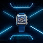 A TAG Heuer Monaco Chronograph wristwatch with a blue strap and silver square case, featuring a skeletonised dial, is centered in a futuristic room lit by blue neon lights.