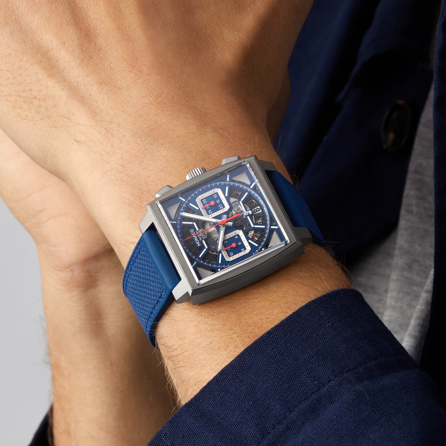 Someone is wearing a TAG Heuer Monaco Chronograph with a square face, blue strap, and skeletonised dial on their wrist.