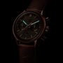 A dark-toned image of the TAG Heuer Special Edition Carrera Chronosprint x Porsche shows its leather strap and chronograph hands, powered by an automatic movement, partially illuminated with a green glow.