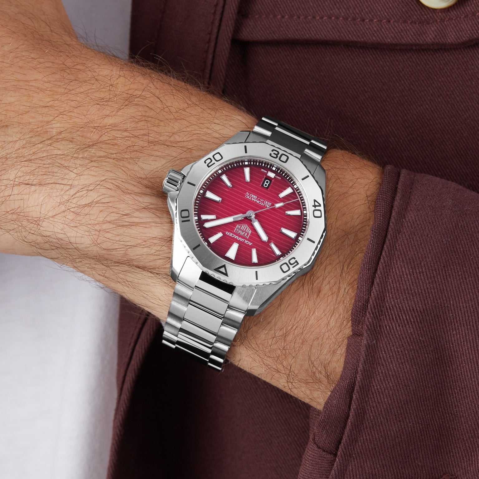 A person wearing a TAG Heuer Aquaracer Professional 200 with a smokey-red sunray brushed dial and silver hour markers, shown from the wrist down against a brown sleeve and stainless steel case.