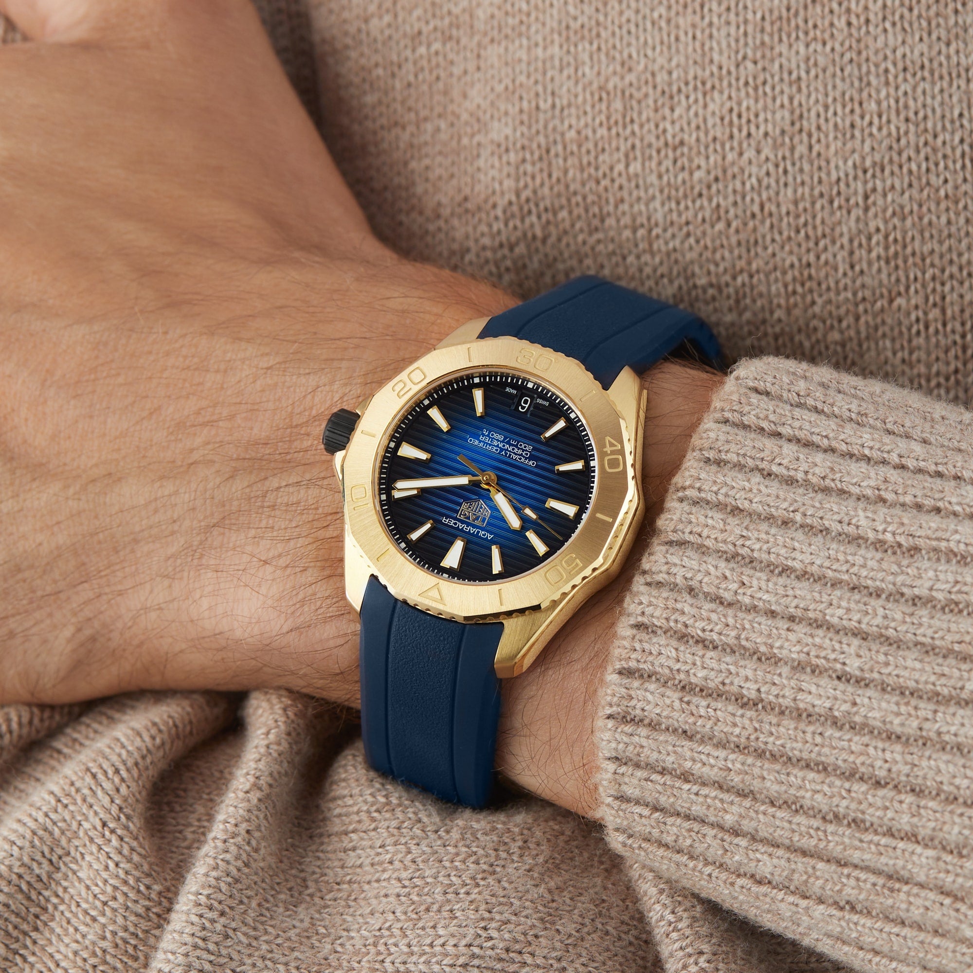 A TAG Heuer Aquaracer Professional 200 Date wristwatch in yellow gold with a blue strap and blue dial with yellow accents, worn on the left wrist with a beige sweater.