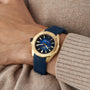A TAG Heuer Aquaracer Professional 200 Date wristwatch in yellow gold with a blue strap and blue dial with yellow accents, worn on the left wrist with a beige sweater.