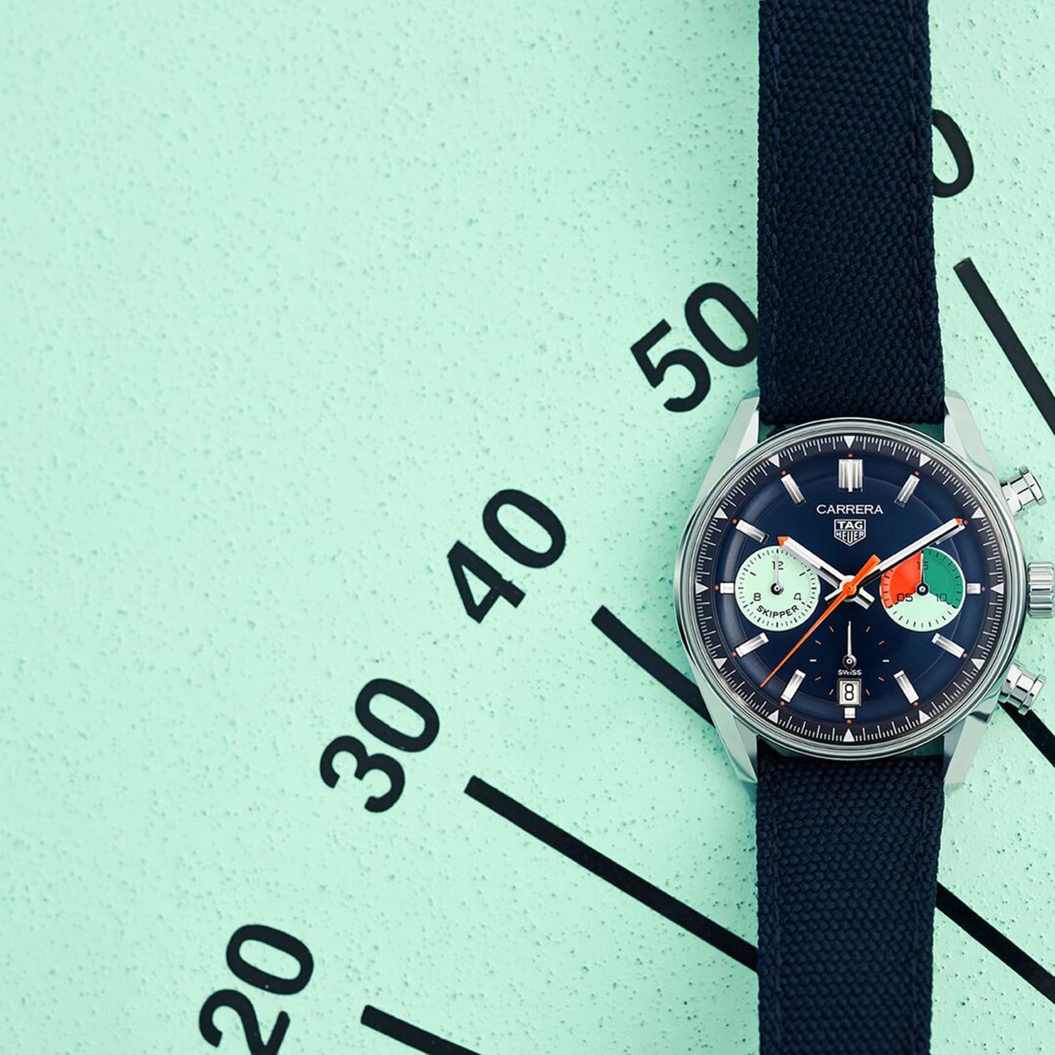 A TAG Heuer Carrera Chronograph Skipper wristwatch with a black strap rests diagonally on a mint green surface, its elegant dial marked in ten-minute increments—a tribute to the racing heritage of the Carrera brand.