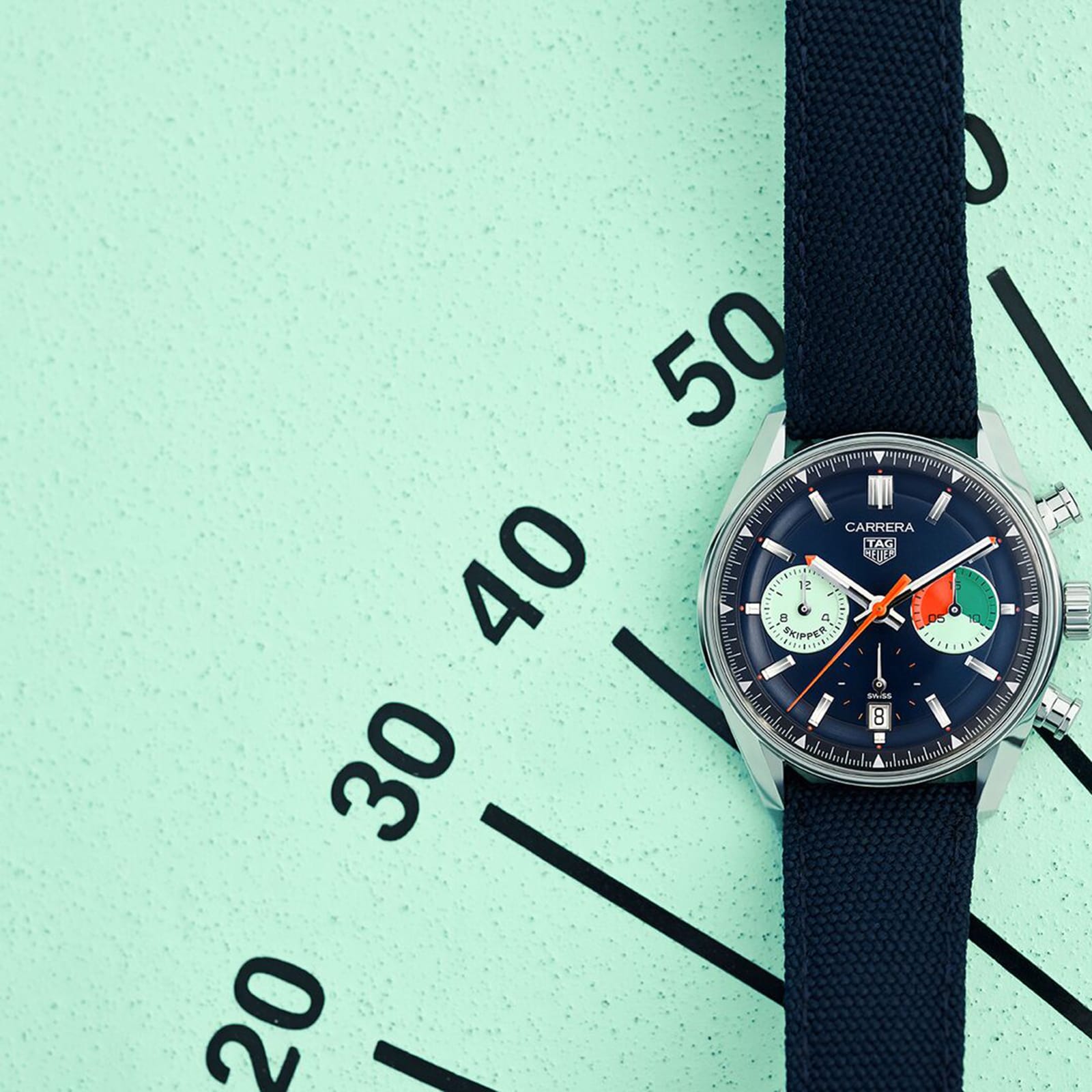 A TAG Heuer Carrera Chronograph Skipper wristwatch with a black strap rests diagonally on a mint green surface, its elegant dial marked in ten-minute increments—a tribute to the racing heritage of the Carrera brand.